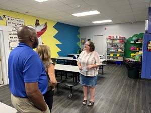one female stands in the center of a room giving a tour of the rock house kids facility. one male and one female are seen on the left side of frame watching the woman talk about rock house kids.