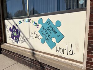 painted mural on an exterior wall. white background with two puzzle pieces. text says Rock House Kids and a quote about providing safe refuge for children.